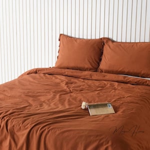 以下が含まれることがあります： さび色の羽毛布団カバーと、それに合わせた枕カバー2枚。羽毛布団カバーは柔らかく快適な生地でできており、シンプルでエレガントなデザインです。寝具は白い木製のヘッドボードのあるベッドに置かれています。