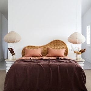May include: A bedroom with a brown wicker headboard, two peach-coloured pillows, and a dark brown linen duvet. Two white pendant lights hang from the ceiling, and there are two small white side tables with plants on them.