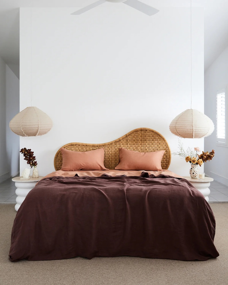 May include: A bedroom with a brown wicker headboard, two peach-coloured pillows, and a dark brown linen duvet. Two white pendant lights hang from the ceiling, and there are two small white side tables with plants on them.