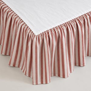 May include: A bed skirt featuring a solid white top platform and a gathered, ruffled drop with vertical stripes in a muted terracotta red and white color palette. The fabric has a textured appearance, and the skirt is designed to drape around the base.