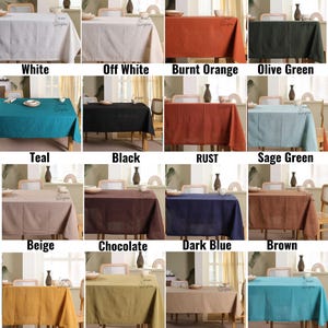 Op de afbeelding: Een verzameling rechthoekige tafelkleden in verschillende kleuren, waaronder wit, gebroken wit, verbrand oranje, olijfgroen, blauwgroen, zwart, roest, saliegroen, beige, chocolade, donkerblauw, bruin, kurkuma, mosgroen, kameel en blauwgroen. Elk tafelkleed wordt op een houten tafel gepresenteerd.