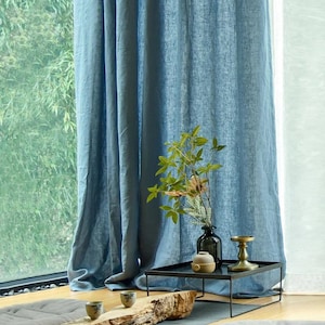 May include: A pair of blue linen curtains hang in a window, with a small wooden table and a vase of greenery in front of them.