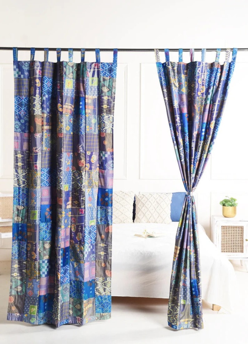 K&ouml;nnte beinhalten: Zwei blaue und violette Patchwork-Vorh&auml;nge mit geometrischem Muster. Die Vorh&auml;nge sind an einer Metallstange aufgeh&auml;ngt und mit Stoffb&auml;ndern zur&uuml;ckgebunden.