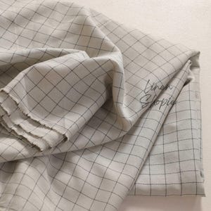 以下が含まれることがあります： 白と黒の格子模様の入った薄いグレーのリネン生地のクローズアップ。生地は折り畳まれ、ドレープされており、素材の質感と織り方を示しています。