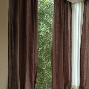 May include: Two sets of brown linen curtains hang in front of a window with a view of green trees. The curtains are pulled back to reveal a view of the trees outside.