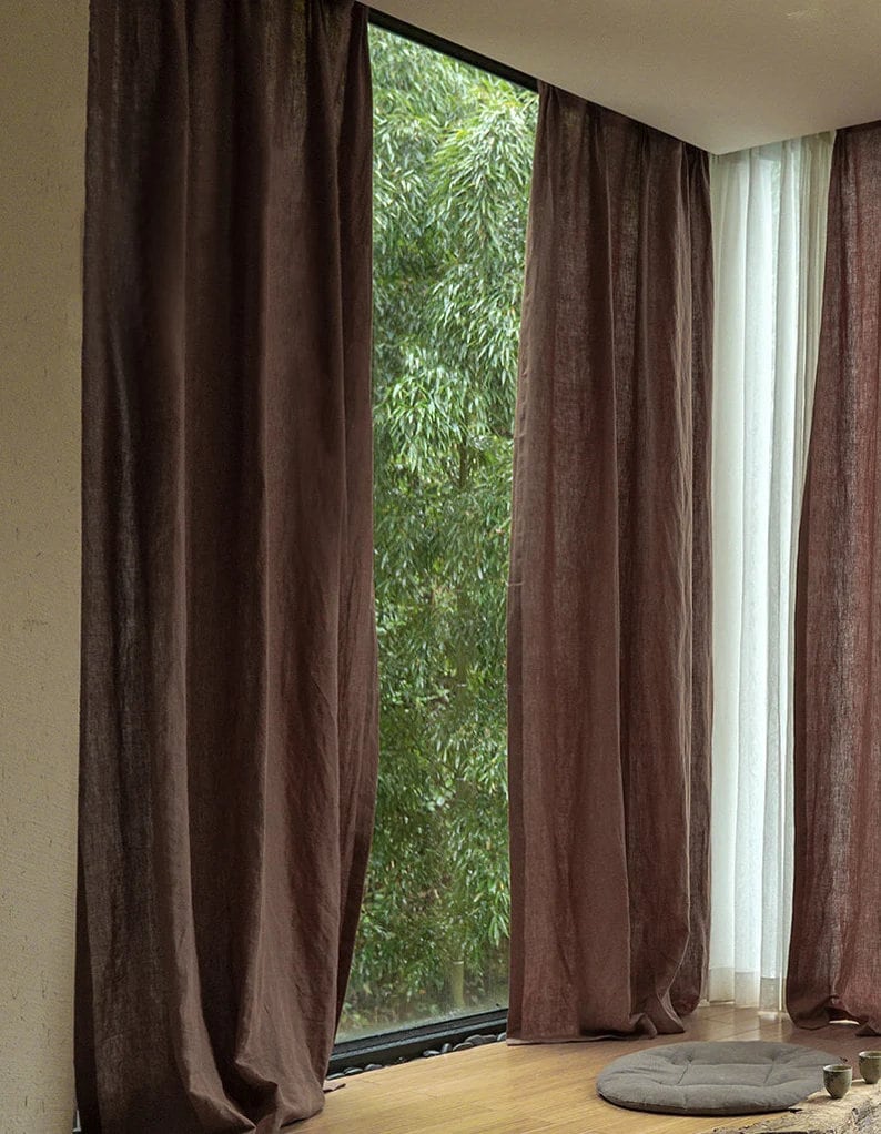 May include: Two sets of brown linen curtains hang in front of a window with a view of green trees. The curtains are pulled back to reveal a view of the trees outside.