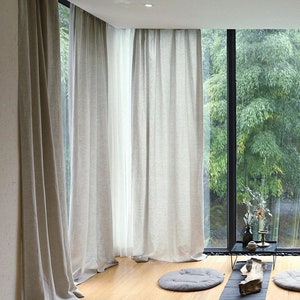 May include: A room with large windows and white linen curtains. The windows overlook a lush green garden. The floor is made of light wood and there are three gray cushions on the floor.