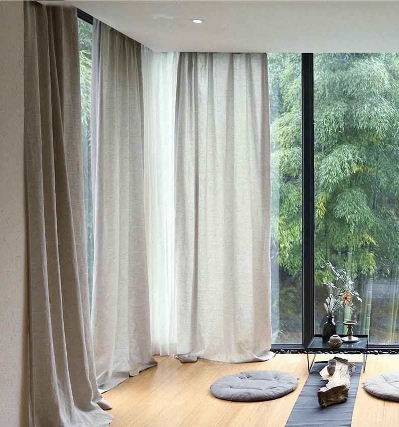 May include: A room with large windows and white linen curtains. The windows overlook a lush green garden. The floor is made of light wood and there are three gray cushions on the floor.