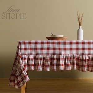 Könnte beinhalten: Eine rot-weiß karierte Tischdecke mit gerüschtem Rand. Die Tischdecke liegt auf einem Holztisch, mit einer Holzschale, einer weißen Vase und einem Büschel trockenem Gras. Der Text "Linen SHOPie" befindet sich in der oberen linken Ecke.