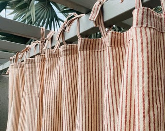May include: Cream-colored curtains with vertical red stripes hang from a gray wooden structure. The curtains are tied to the structure with matching red and cream fabric ties. The background features green foliage.