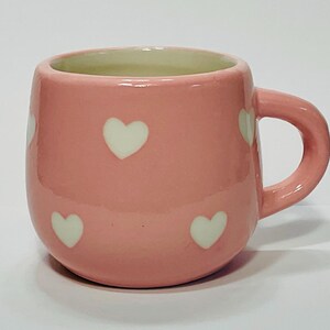 Puede incluir: Una taza de cer&aacute;mica rosa con un patr&oacute;n de corazones blancos. La taza tiene forma redondeada, un asa y un interior verde claro. La taza mide aproximadamente 7.6 cm de alto y 7.6 cm de di&aacute;metro.