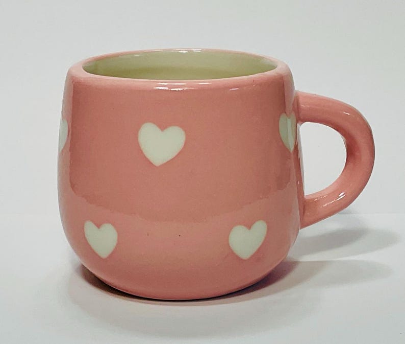 Puede incluir: Una taza de cer&aacute;mica rosa con un patr&oacute;n de corazones blancos. La taza tiene forma redondeada, un asa y un interior verde claro. La taza mide aproximadamente 7.6 cm de alto y 7.6 cm de di&aacute;metro.
