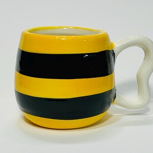 Puede incluir: Una taza de cer&aacute;mica con un dise&ntilde;o de rayas amarillas y negras, que se asemeja a una abeja. La taza tiene un asa blanca y curvada. El interior de la taza es blanco.