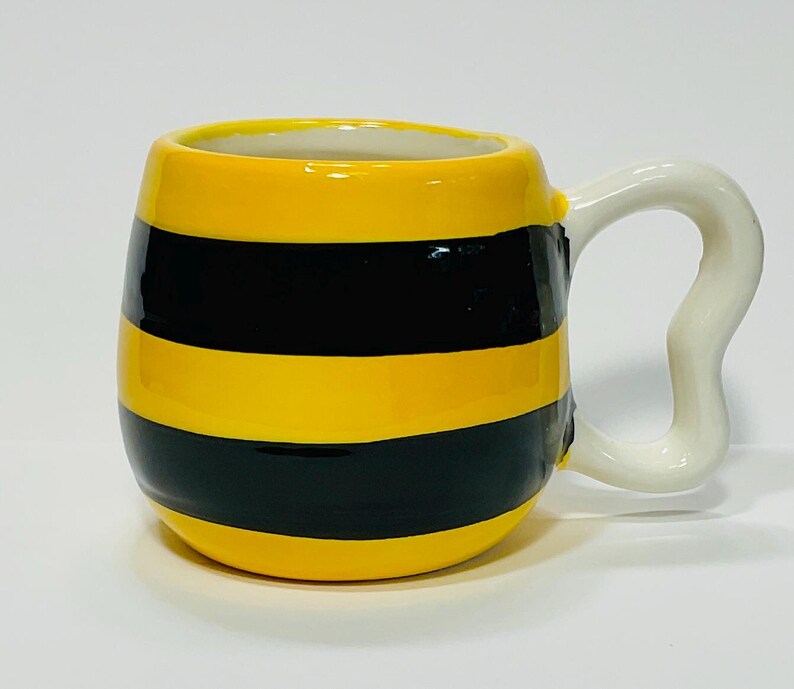 Puede incluir: Una taza de cer&aacute;mica con un dise&ntilde;o de rayas amarillas y negras, que se asemeja a una abeja. La taza tiene un asa blanca y curvada. El interior de la taza es blanco.