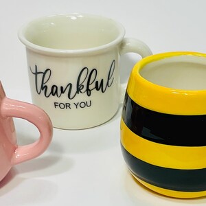Puede incluir: Tres tazas de cer&aacute;mica. Una es rosa con corazones blancos, otra es blanca con "thankful FOR YOU" en escritura negra, y la tercera es amarilla con rayas negras, que se asemeja a una abeja.