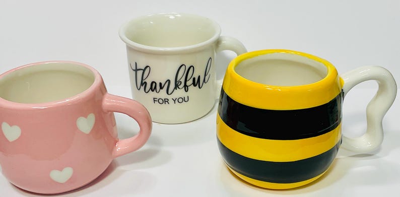 Puede incluir: Tres tazas de cer&aacute;mica. Una es rosa con corazones blancos, otra es blanca con "thankful FOR YOU" en escritura negra, y la tercera es amarilla con rayas negras, que se asemeja a una abeja.