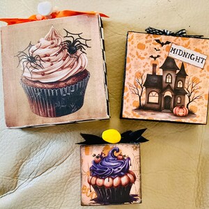 May include: Three Halloween-themed decorative boxes. One features a cupcake with spiders, another a haunted house with the word "MIDNIGHT", and the third a cupcake with purple frosting. Each box has a ribbon and decorative accents.