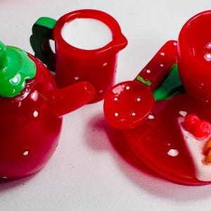 May include: A strawberry-themed tea set with a teapot, creamer, cup and saucer, and a small plate with a strawberry-shaped cake. The set is made of red and green plastic.