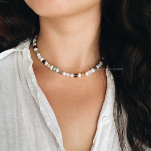 Puede incluir: Un collar de cuentas multicolores con perlas, que presenta una variedad de piedras, incluyendo turquesa, amatista y cornalina. El collar lo lleva una persona con el pelo largo y oscuro y una camisa blanca.