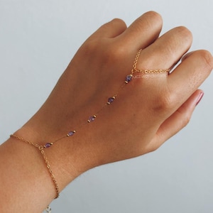 Pode incluir: Pulseira de corrente de mão dourada com pequenas contas roxas. A pulseira conecta-se a um anel e envolve o pulso. O design inclui uma corrente delicada e pequenas pedras preciosas roxas.