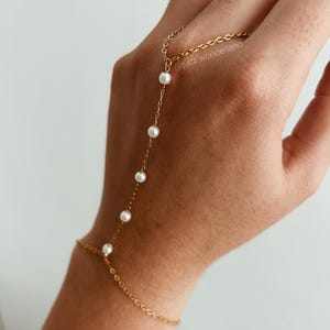 Puede incluir: Un brazalete de cadena dorada con un detalle de perla que se extiende hasta la mano y forma un anillo en el dedo. El brazalete se lleva en la mano de una persona.  @jucz_wonderland