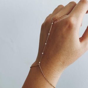 Può includere: Un braccialetto a catena dorata con piccole perle bianche, collegato a un anello che può essere indossato al dito. Il braccialetto è indossato al polso. Il testo '@juczwonderland' è visibile sull'anello.