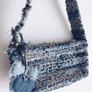 Pode incluir: Uma bolsa de ombro de ganga feita à mão com uma textura tecida. A bolsa é predominantemente azul com retalhos de ganga mais claros e escuros e bordas desfiadas. A alça também é feita de ganga tecida.