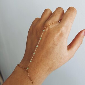 Puede incluir: Un brazalete de cadena dorada con una cadena delicada y pequeñas cuentas verdes. El brazalete está diseñado para usarse en la muñeca y la mano, con la cadena extendiéndose hacia arriba del brazo.