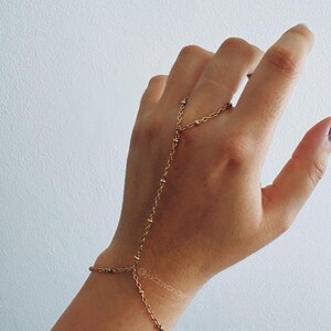 Puede incluir: Una pulsera de mano dorada que presenta una delicada cadena de eslabones con pequeñas cuentas espaciadas uniformemente. La pieza de joyería conecta una pulsera de muñeca con un anillo que rodea el dedo medio, mostrando el texto @juczwonderland.