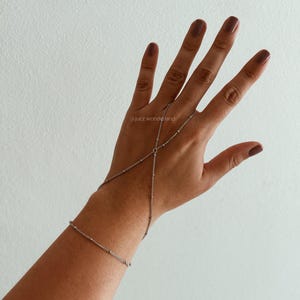 Puede incluir: Un brazalete de cadena de plata con un diseño delicado. El brazalete se lleva en la muñeca y se extiende por el brazo, conectándose a un anillo en el dedo índice. La cadena tiene pequeñas cuentas redondas.