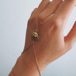 Puede incluir: Pulsera de cadena de mano dorada con un colgante de sol con una cara sonriente. La pulsera se conecta a un anillo para el dedo y se envuelve alrededor de la muñeca. La joya se muestra sobre un fondo claro.