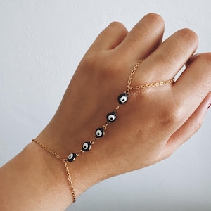 Pode incluir: Uma pulseira de corrente de mão dourada com uma série de amuletos de olho grego em preto e branco. A pulseira conecta-se a um anel e envolve o pulso. O fundo é uma superfície branca lisa.