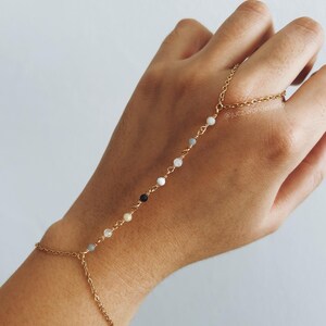 Pode incluir: Um bracelete de corrente dourada com pequenas contas brancas, cinzas e pretas. O bracelete tem uma corrente longa que se estende pelo braço e envolve o dedo.