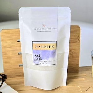 Peut inclure: Pochette blanche avec une étiquette qui indique "The Fine Feet Company" et "Nannies Bath Soak with Coconut & Lavender Oil Relax".