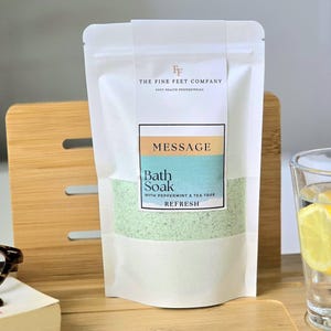 Peut inclure: Une pochette blanche contenant un bain moussant vert. La pochette porte une étiquette indiquant "The Fine Feet Company" et "Message Bath Soak with Peppermint & Tea Tree Refresh".