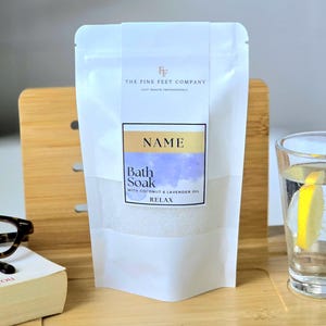 Peut inclure: Pochette blanche avec une étiquette qui dit "THE FINE FEET COMPANY" et "NAME Bath Soak WITH COCONUT & LAVENDER OIL RELAX". La pochette contient des sels de bain blancs.