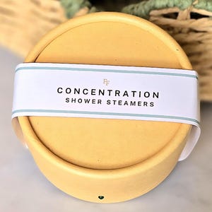 May include: A circular, yellow shower steamer container with a white band displaying the words "CONCENTRATION SHOWER STEAMERS" in black. The band has a light blue border. The background is blurred.