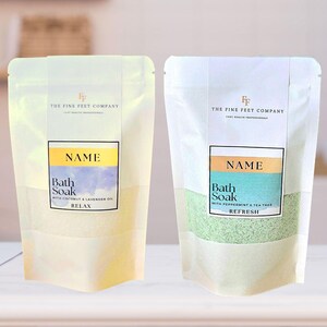 Peut inclure: Deux sachets blancs avec des étiquettes indiquant "The Fine Feet Company". Le sachet de gauche a un fond jaune clair et contient un bain moussant blanc avec le texte "Bath Soak with Coconut & Lavender Oil Relax". Le sachet de droite a un fond blanc et contient un bain moussant vert avec le texte "Bath Soak with Peppermint & Tea Tree Refresh".