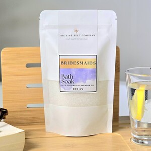 Peut inclure: Un sachet blanc de bain moussant avec le texte "BRIDESMAIDS" et "Bath Soak" à l'huile de noix de coco et de lavande. Le sachet est sur un plateau en bois avec un verre d'eau et une tranche de citron.