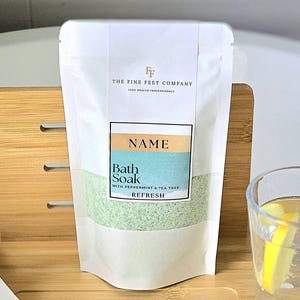 May include: White pouch with a green bath soak inside. The pouch has the text "THE FINE FEET COMPANY" and "NAME" on it. The bath soak is labeled "Bath Soak with Peppermint & Tea Tree Refresh".