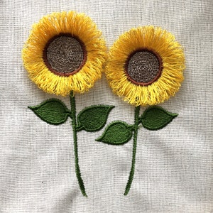 May include: Two embroidered sunflowers with green stems and leaves on a white linen background.