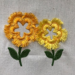 May include: Two yellow embroidered flowers with white centers and green stems on a white fabric background.