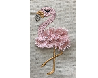 May include: An embroidered patch on a beige fabric background featuring a flamingo. The flamingo has a pink stitched head with a closed eye, a gold and silver beak, and a body covered in layered pink fringe. It has two thin gold legs.
