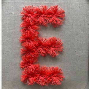 May include: A large letter E made of red yarn, with each loop of the letter formed by a cluster of yarn pom-poms. The letter is displayed on a light gray linen background.