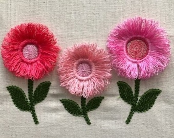 May include: Three embroidered flowers with pink, light pink, and red petals. The flowers are stitched on a white fabric background.