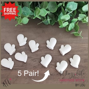 May include: A collection of ten small, white mitten-shaped ornaments arranged on a wooden surface. The text "5 Pair!" is displayed with an arrow pointing to the mittens. Green foliage is visible in the background. The text "All things little handmade with love by Lou" is also visible.