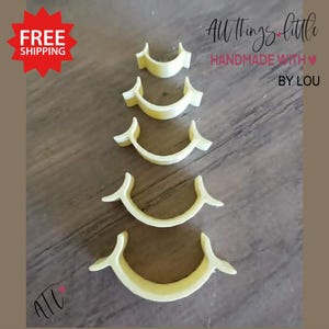 May include: Four yellow, crescent-shaped cookie cutters of varying sizes are arranged vertically. The cutters have a curved base with pointed tips on each end. The image includes the text "All Things Little Handmade With by Lou".