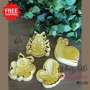 May include: Four yellow plastic cookie cutters in the shapes of a ladybug, snail, bee, and butterfly. The ladybug has a dotted design, and the snail has a spiral shell. The bee and butterfly have detailed wing designs. The text "All things little handmade with love by Lou" is visible.