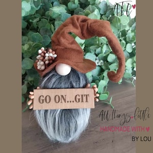 May include: A brown gnome with a long brown hat and a gray beard is holding a sign that says "GO ON...GIT".