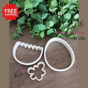 May include: Three white plastic cookie cutters in the shapes of an egg, a flower, and a cracked eggshell. The background includes green foliage and text that reads "All things little HANDMADE WITH BY. LOU". A red starburst with "FREE SHIPPING" is in the upper left.